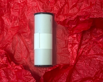 May include: A cylindrical product container with a white body and a black rim, wrapped with a white band. The container is set against a backdrop of crumpled red tissue paper, creating a contrasting visual texture.