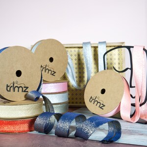 May include: Assortment of spools of ribbon in various colours, including silver, pink, gold, and blue. The ribbon is glittery and has a textured finish. The spools are labelled "trimz".