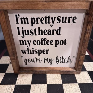 May include: A wooden framed sign with the text "I'm pretty sure I just heard my coffee pot whisper 'you're my bitch'" in black lettering on a white background. The frame is a dark brown color.