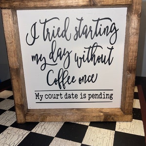 以下が含まれることがあります： 白い背景に黒い筆記体で「I tried starting my day without coffee once. My court date is pending」と書かれた木製の額入りサイン。 フレームは明るい茶色です。