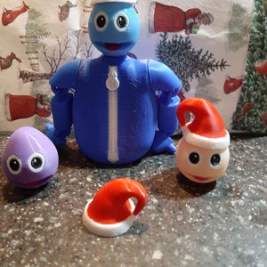 May include: A blue articulated figure wearing a Santa hat, with a matching hat and two other figures with googly eyes. The figure has a white zipper and is posed against a festive background.