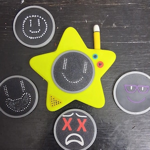 May include: A yellow star-shaped drawing toy with a stylus, surrounded by four black circular drawing pads. Each pad features a different smiley face design, including a happy face, a sad face, and a face with glasses.