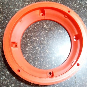 May include: A vibrant orange circular plastic component with a central aperture. It has several evenly spaced holes around its edge, suggesting it's part of a larger assembly. The plastic has a slightly textured surface, and is likely a manufactured part.