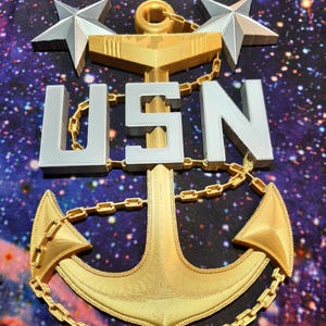 May include: A gold anchor with a chain and two silver stars, with the letters USN in silver.