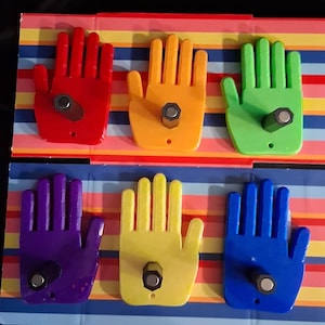 May include: Six colorful hand-shaped wall hooks in red, orange, green, purple, yellow, and blue. Each hook has a metal bolt for hanging and is mounted on a striped background.