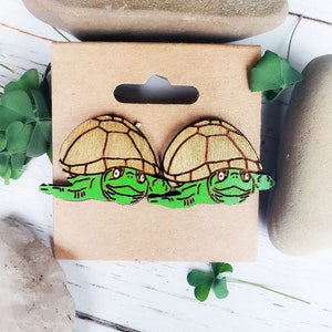 May include: A pair of wooden turtle earrings. The turtles are painted green with brown shells. The earrings are displayed on a brown card.
