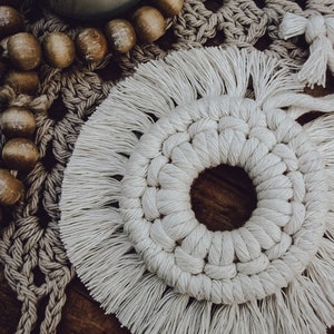 May include: A white macrame coaster with a fringe border. The coaster is round and has a hole in the center. It is sitting on a wooden surface.
