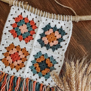 May include: A white crocheted wall hanging with four colorful granny squares. The squares feature a variety of colors, including orange, pink, teal, and mustard yellow. The hanging has a fringe border in matching colors.