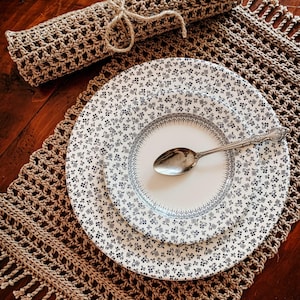 May include: A set of two crocheted placemats with fringe, one rolled up and tied with twine, and a set of two white ceramic plates with a black floral pattern.