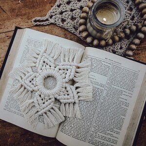 Peut inclure: Une étoile en macramé blanche avec des franges accrochée à un livre ouvert avec du texte. Le livre est posé sur une surface en bois avec une bougie et des perles.
