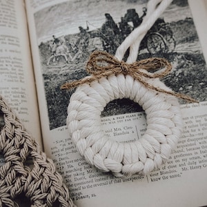 May include: A white crocheted wreath with a brown twine bow. The wreath is hanging from a book with the text "ROAD, A PALE FLARE ON IT WAS YET FAR OFF."