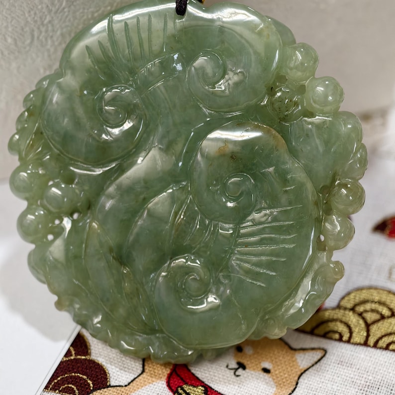 May include: A light green jade pendant with intricate carvings. The pendant features a swirling design with floral and abstract elements. The jade has a translucent quality, and a black cord is attached for wearing.