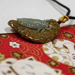 May include: A jade pendant featuring a detailed cicada resting on a leaf-shaped base. The cicada is a translucent green, while the leaf is a mottled brown. The pendant hangs from a black cord with a gold-colored knot. The background is a red fabric with white and gold patterns.
