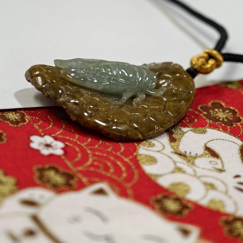 May include: A jade pendant featuring a detailed cicada resting on a leaf-shaped base. The cicada is a translucent green, while the leaf is a mottled brown. The pendant hangs from a black cord with a gold-colored knot. The background is a red fabric with white and gold patterns.