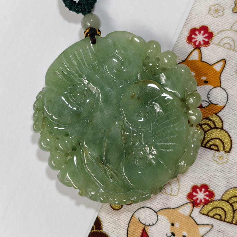 May include: A carved jade pendant with a green hue, featuring a detailed floral and fish design. The pendant is suspended from a dark green cord with a bead. The background includes a patterned fabric with cartoon foxes and flowers.