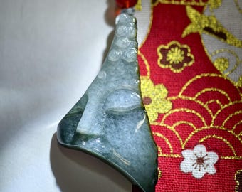 May include: A jade pendant carved with a serene Buddha face. The pendant is a mix of green and white hues, with a smooth, polished finish. It hangs from a string with a red bead. The background features a red fabric with gold floral patterns.