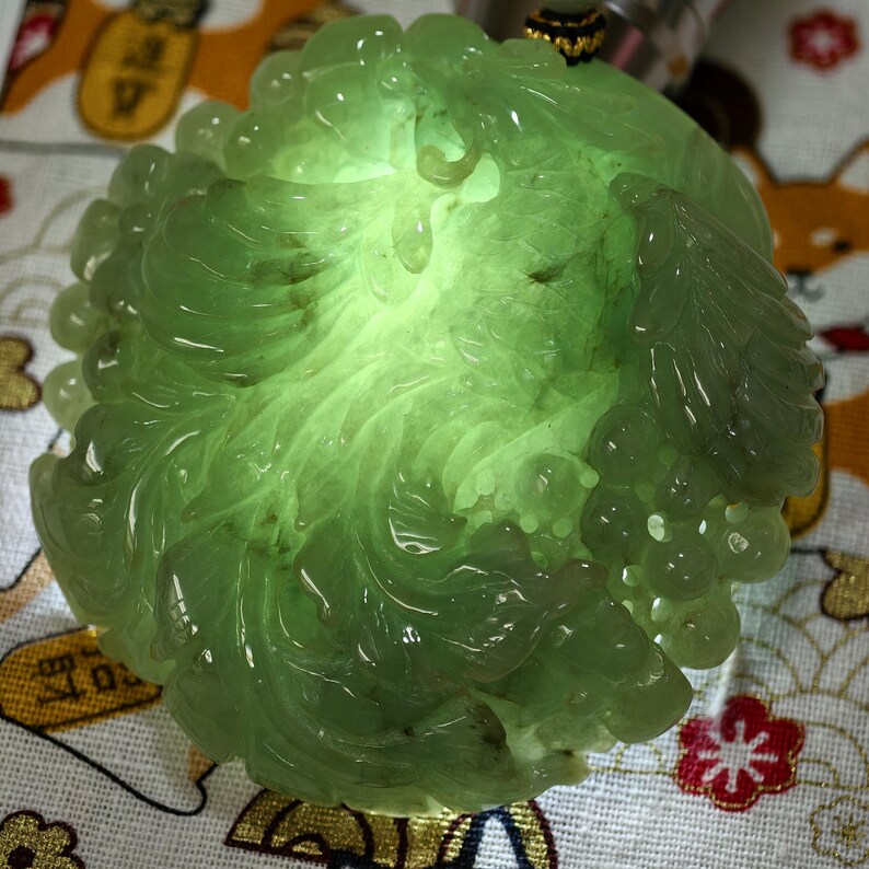 May include: A translucent, light green jade pendant carved in a floral design. The pendant has a detailed, swirling pattern with a smooth, polished finish. The jade glows with an internal light, highlighting the intricate details of the carving.