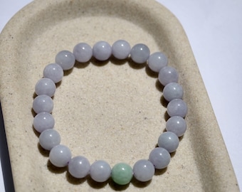 May include: A beaded bracelet featuring round, light purple beads with one green bead. The bracelet is displayed on a beige, textured surface. The beads appear to be made of a smooth, polished material.