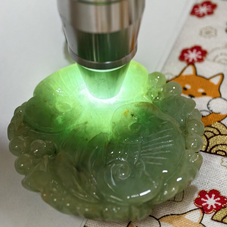 May include: A carved jade pendant in shades of green, illuminated by a bright light. The pendant features a detailed relief design of a fish and floral elements. The jade is polished and has a translucent quality.