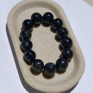 May include: A black beaded bracelet with a glossy finish. The bracelet is made of round, dark beads, likely gemstones, arranged in a circular shape. The bracelet is displayed on a light beige, oval-shaped tray.
