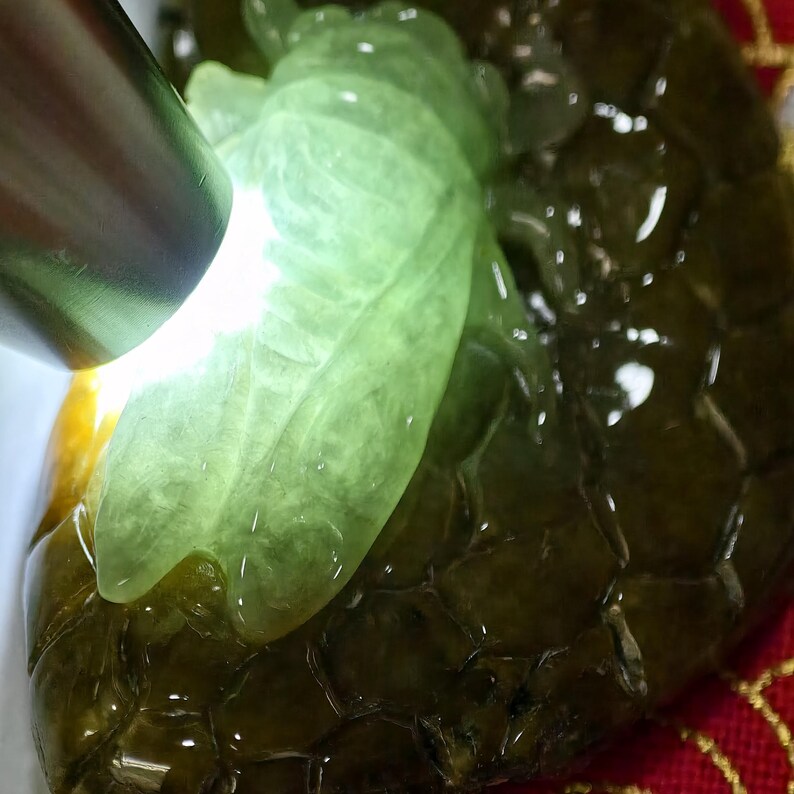 May include: Close-up of a jade carving with a leaf design. The jade is a translucent light green color, with darker green and brown tones in the background. A bright light shines on the jade, highlighting its details.