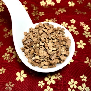 Puede incluir: Una cuchara de cerámica blanca llena de astillas de madera marrón. La cuchara está sobre un fondo rojo con un patrón floral dorado.