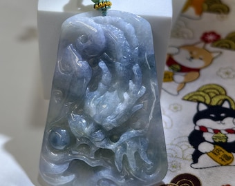 May include: A rectangular jade pendant with a carved dragon and fish design. The pendant is primarily gray and blue with a green and gold accent at the top. The pendant is suspended from a green and white cord.