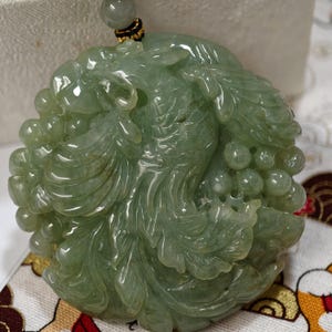 May include: A jade pendant carved with a phoenix design. The pendant is a light green color and features intricate details of the bird's feathers and body. The pendant is suspended from a black cord with a small bead.