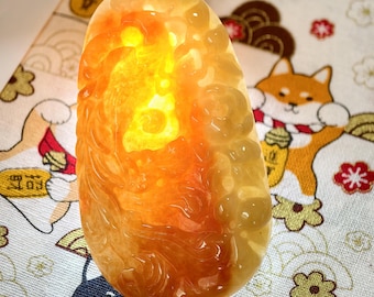 May include: A translucent, oblong pendant with a warm, amber and orange color gradient. The pendant has a carved design and is suspended by a dark cord. The background features a patterned fabric with cartoon illustrations.