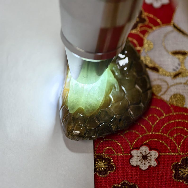 May include: A close-up of a dark green, translucent gemstone illuminated by a flashlight. The gemstone has a textured surface and a glowing, light green interior. The background includes a red and gold patterned fabric.