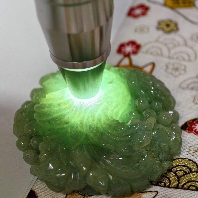 May include: A light green jade carving, illuminated by a bright light. The jade is carved in a floral design with rounded edges. The light source is a silver-colored tool.