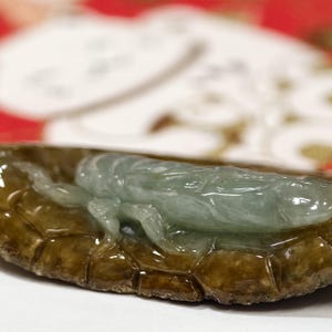 May include: A jade carving of a grasshopper. The insect is light green and rests on a brown, textured base. The carving is detailed, with visible legs and body segments. The background is blurred, with red and white elements.