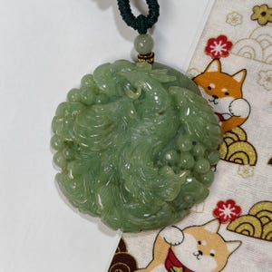 May include: A jade pendant with a carved dragon design. The green pendant is round and has a dark green cord. The pendant is displayed on a white surface with a patterned fabric.