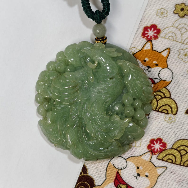 May include: A jade pendant with a carved dragon design. The green pendant is round and has a dark green cord. The pendant is displayed on a white surface with a patterned fabric.