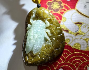 May include: A jade pendant featuring a light green cicada carved on a larger, olive-green stone. The pendant is suspended from a black cord with a gold-colored accent. The background includes a red fabric with floral and geometric patterns.