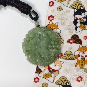 May include: A light green jade pendant with intricate carvings, suspended from a black and green cord. The pendant is round and features a detailed floral design. The background includes a patterned fabric with cartoon Shiba Inu dogs and Japanese motifs.