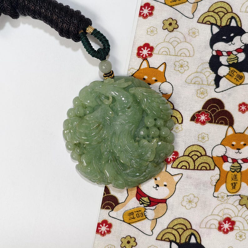 May include: A light green jade pendant with intricate carvings, suspended from a black and green cord. The pendant is round and features a detailed floral design. The background includes a patterned fabric with cartoon Shiba Inu dogs and Japanese motifs.