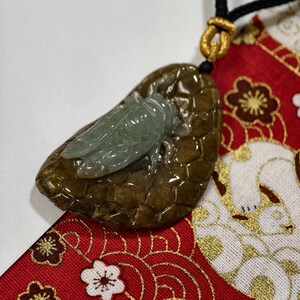 May include: A jade pendant featuring a carved cicada on a brown, textured base. The cicada is a pale green color, and the base has a glossy finish. The pendant hangs from a black cord with a gold-colored clasp.