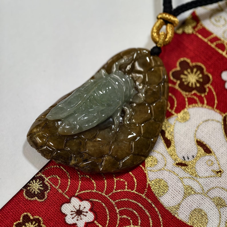 May include: A jade pendant featuring a carved cicada on a brown, textured base. The cicada is a pale green color, and the base has a glossy finish. The pendant hangs from a black cord with a gold-colored clasp.