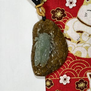 May include: A jade pendant featuring a detailed cicada design. The pendant has a brown, textured background and a light green cicada. The pendant hangs from a black cord with gold accents.