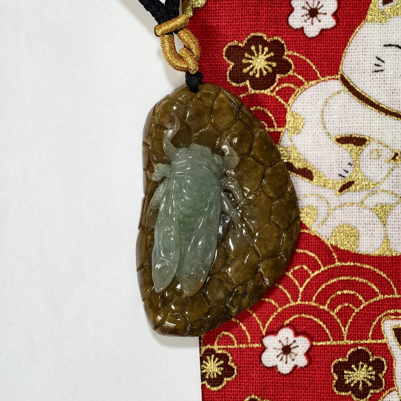 May include: A jade pendant featuring a detailed cicada design. The pendant has a brown, textured background and a light green cicada. The pendant hangs from a black cord with gold accents.