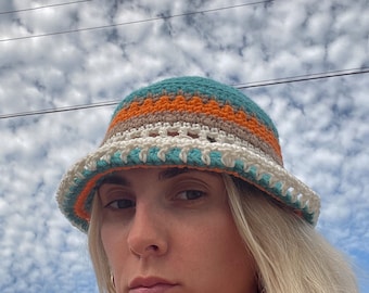 May include: A crocheted bucket hat with teal, orange, white and brown stripes. The hat is worn by a person with blonde hair against a cloudy sky.