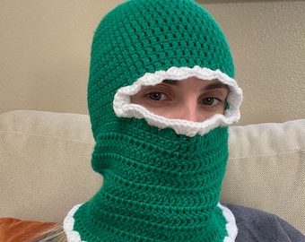May include: A green crocheted balaclava with a white scalloped trim around the face and neck. The handmade head covering is made of textured yarn and is designed to cover the head and neck.