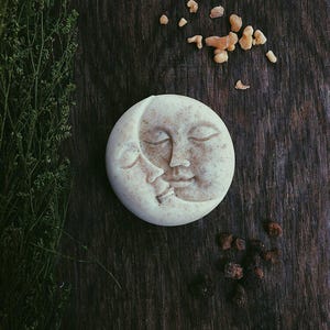 May include: A white soap bar shaped like a crescent moon with a face sculpted into the surface. The soap is on a dark brown wooden surface with small pieces of brown and orange resin scattered around it.