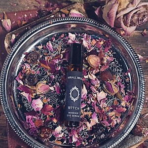 Könnte beinhalten: Eine kleine Glasflasche Mirall Bru Witch Rosehip Eye Serum liegt auf einem silbernen Tablett mit getrockneten Rosenblättern. Die Flasche hat ein schwarzes Etikett mit weißem Text und einem dekorativen Emblem. Der Hintergrund ist eine Holzoberfläche.