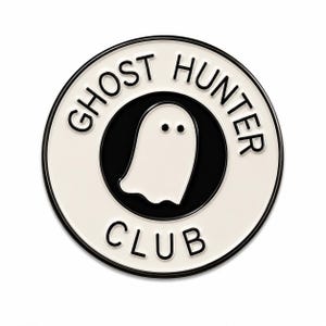 Pode incluir: Um broche de esmalte circular com um centro preto e uma ilustração de um fantasma branco. A borda externa é branca com o texto preto GHOST HUNTER CLUB impresso em disposição circular. O broche tem uma borda fina de metal preto.