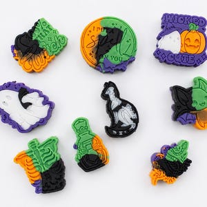 May include: A collection of nine colourful Halloween-themed shoe charms in purple, green, orange, and black. Designs include a ghost, a skeleton, a potion bottle, a cat, and text reading 100% That Witch, I Put A Spell On You, and Wicked Cute.