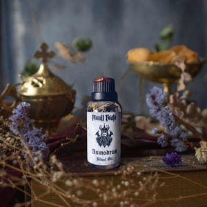 Może przedstawiać: Mała, ciemna butelka szklana z czarną etykietą i białym obrazkiem przedstawiającym koronowaną postać z brodą. Etykieta zawiera napis "Minall Brujo Asmodeus Ritual Oil".