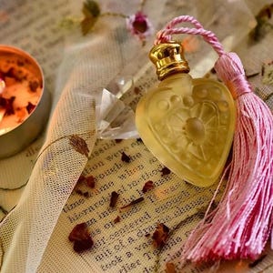 May include: A heart-shaped glass bottle with a gold cap and a pink tassel. The bottle is filled with a clear liquid and has a decorative design on the front. The bottle is sitting on a piece of lace fabric with a candle and other decorative items.
