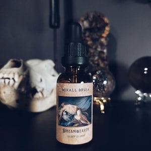 Könnte beinhalten: Eine braune Glasflasche mit schwarzem Tropfer und einem Etikett mit der Aufschrift "Mirall Bruja Dreamweaver Sleep Elixir". Das Etikett zeigt eine Vintage-Illustration einer Frau in einem blauen Kleid.